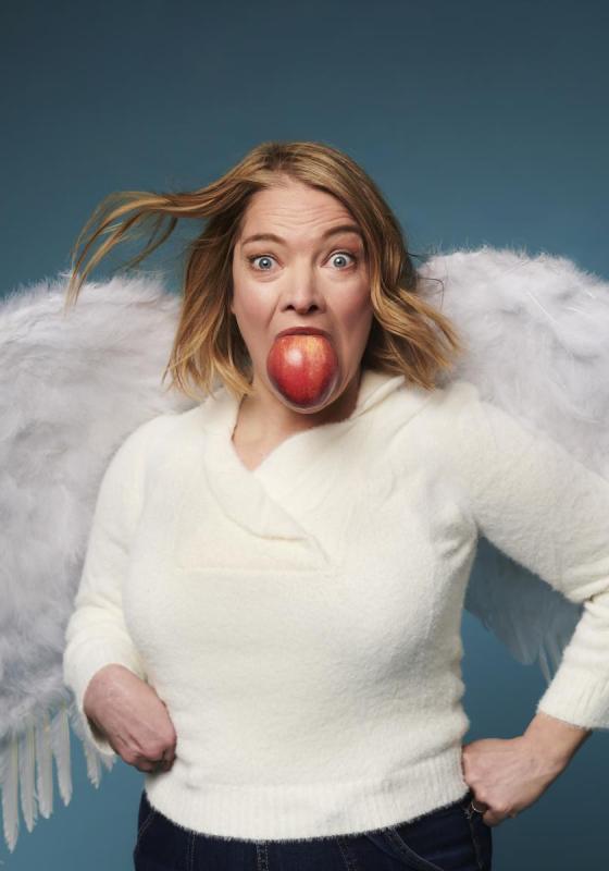 The performer wears fluffy angel wings and has an apple in their mouth as they look at the camera, startled, in front of a sky blue background. 