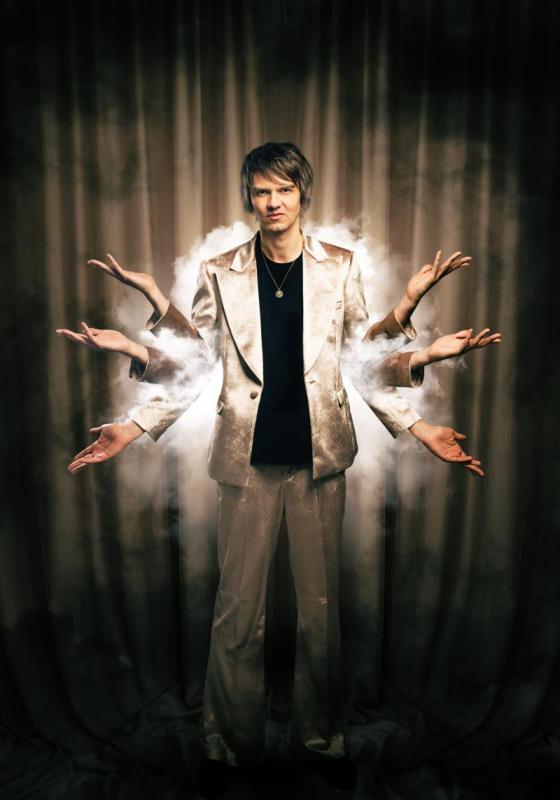 A performer stands in front of a dark curtained background. They are holding up 6 arms, and there is a haze of smoke around their torso. They are wearing a fabulous silvery suit.