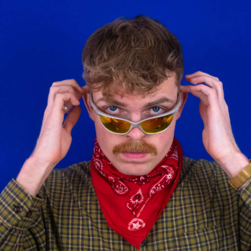 Horatio squares up to a blue background wearing a yellow checked shirt, red bandana around their neck and holding a pair of retro sunglasses halfway down their nose.
