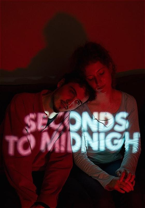 A boy and a girl sit next to each other with the words 'seconds to midnight' projected across their bodies.