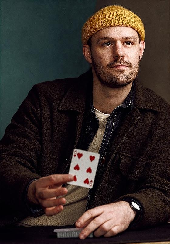 Andrew sits at a table, looking off camera and holding the six of hearts.