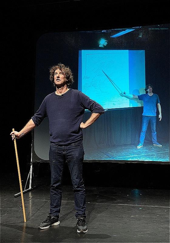 The performer stands holding a long stick in front of a projected screen.