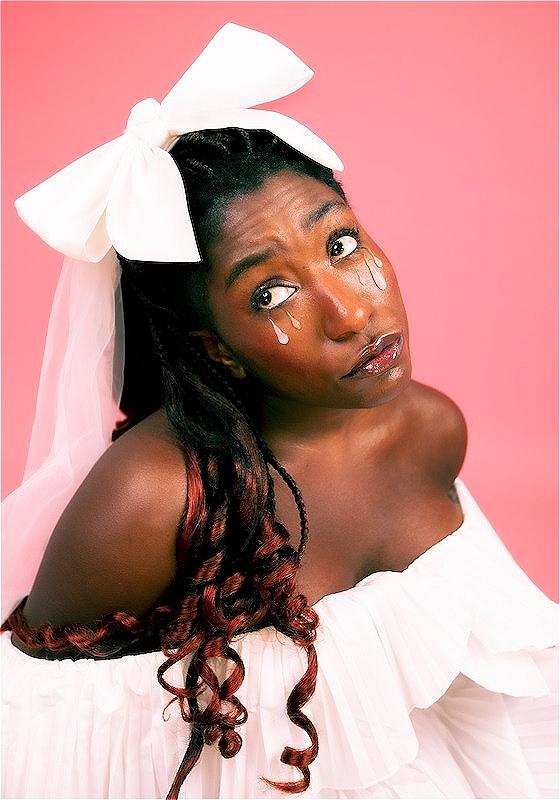 The performer is in front of a pink background with a white bow in her hair and white teardrops drawn on her face