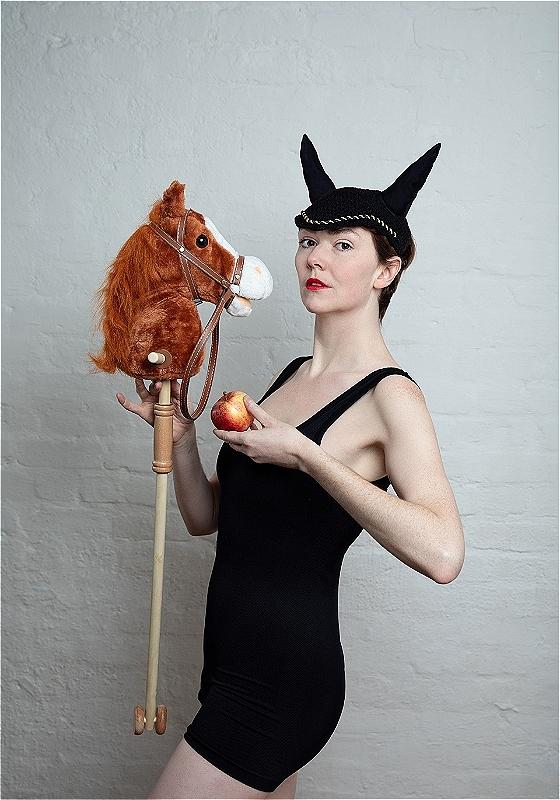 The performer wears black horns and looks to the camera holding an apple and a toy horse.