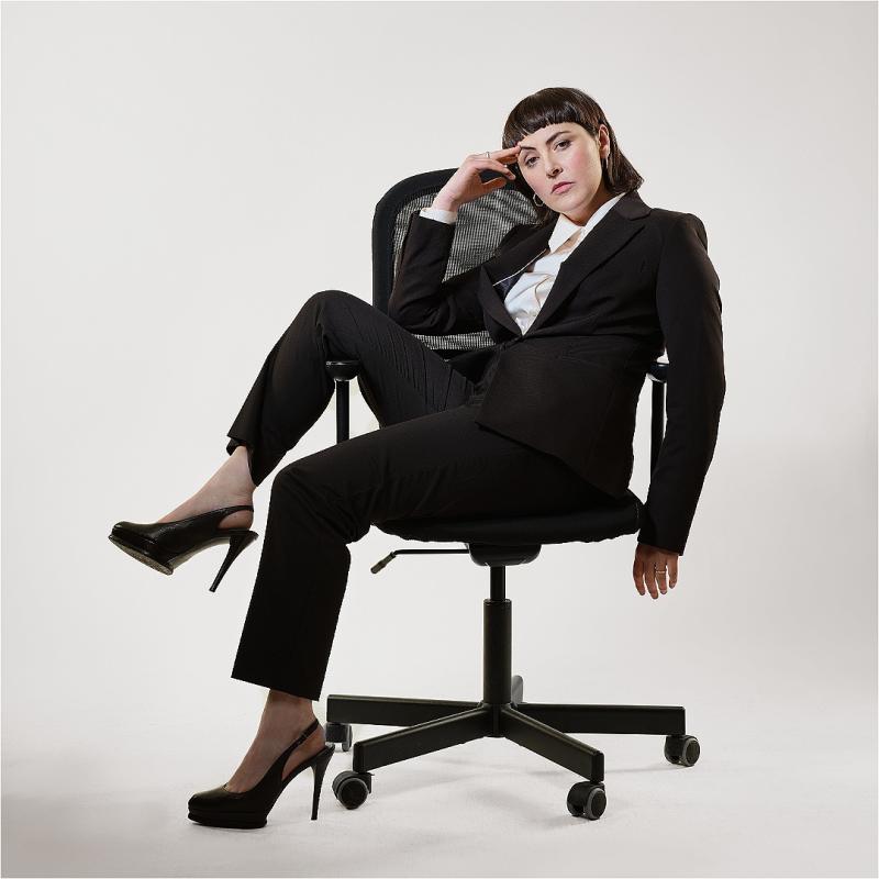 The performer sits in a desk chair wearing a black suit and heels.