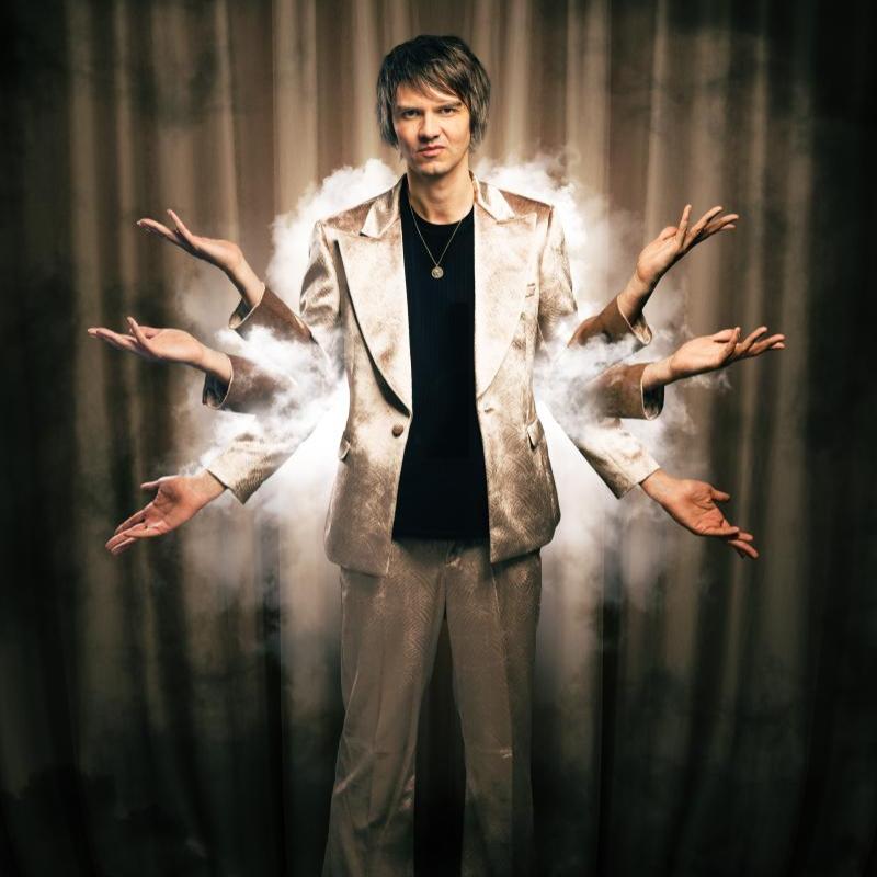 A performer stands in front of a dark curtained background. They are holding up 6 arms, and there is a haze of smoke around their torso. They are wearing a fabulous silvery suit.