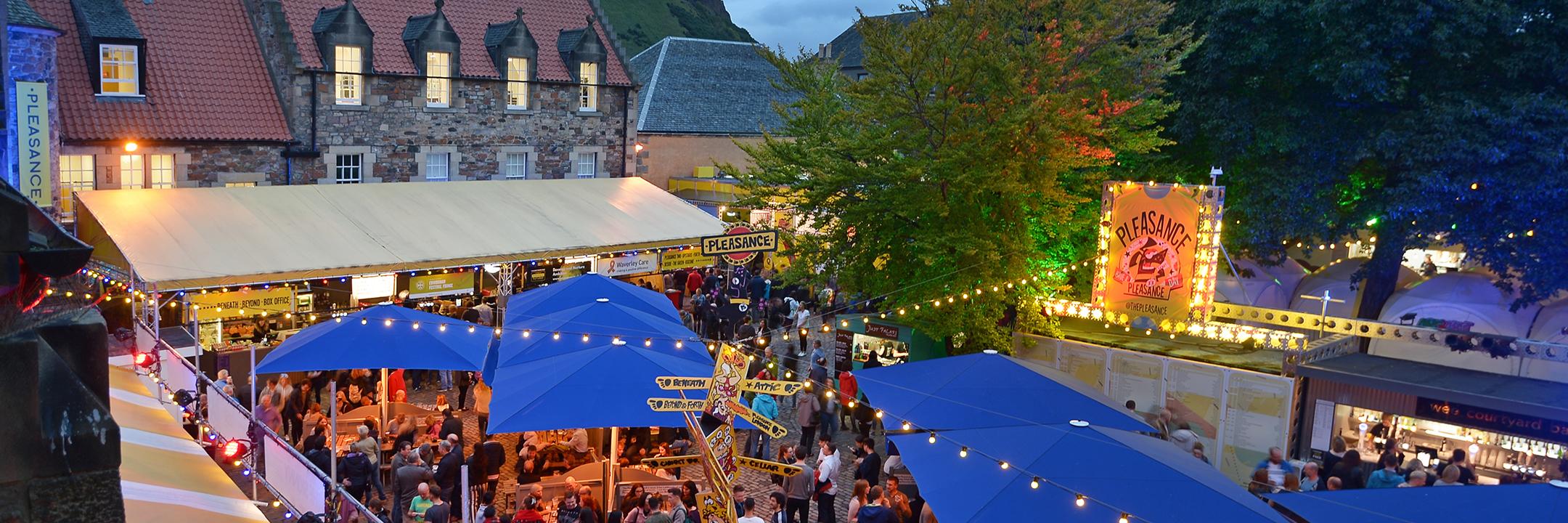 Photo overlooking the Pleasance Courtyard with festoon lights, blue parasols and yellow stretch tents. Scores of audience are glimpsed underneath the canopy.