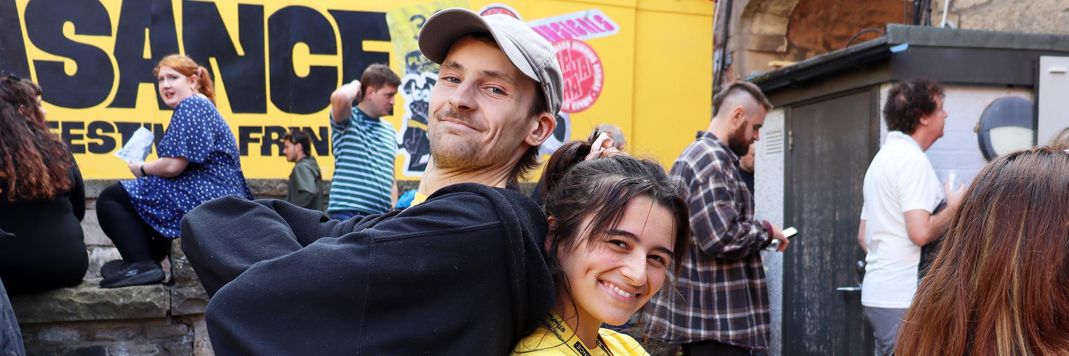 Two smiling volunteers stand back to back in front of the Pleasance's branded wall within the entry to the Pleasance Courtyard.