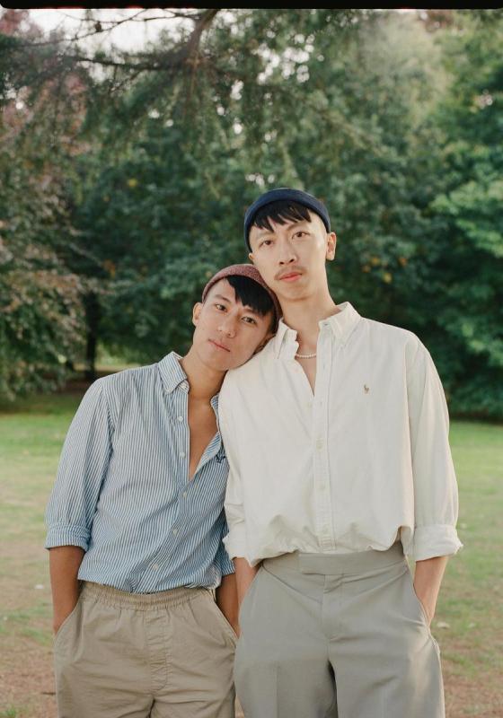 2 People standing in in a park. The shorter person leans their head on the shoulder of the other. They both have their hands in their pockets and are wearing shirts and smart trousers.