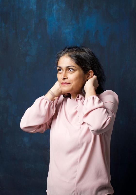 A woman in a pink shirt stands in front of a dark ocean blue backdrop putting her hair behind her ears looking up positively.