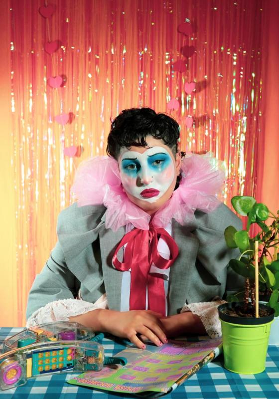Saffia, a non-binary white person with short curly dark hair, wears a pink clown collar with a large red bow and a grey blazer. They are sitting with their arms on a blue gingham table, showing a plant, a noughties magazine and a transparent telephone with multicoloured keys. The backdrop is sunset orange with iridescent fringe and hanging heart garlands.  
