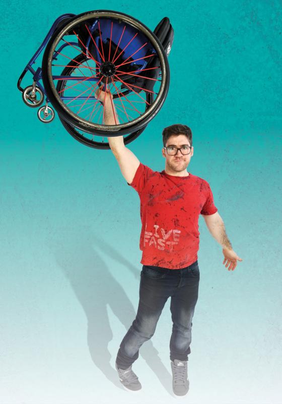 Aaron Simmonds stands against a teal background fading into white. He holds aloft a wheelchair above his head.
