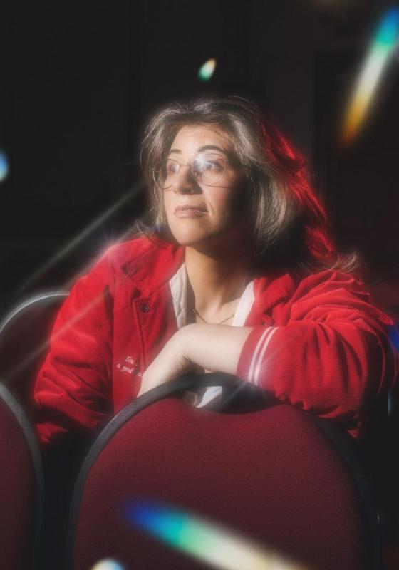 Wearing a red sports jacket and white top, Zoe sits in a theatre with lights reflecting and looks off camera.