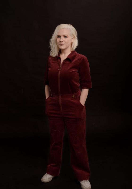 Lulu stands with her hands in her pockets wearing a red velvet jumpsuit against a black background