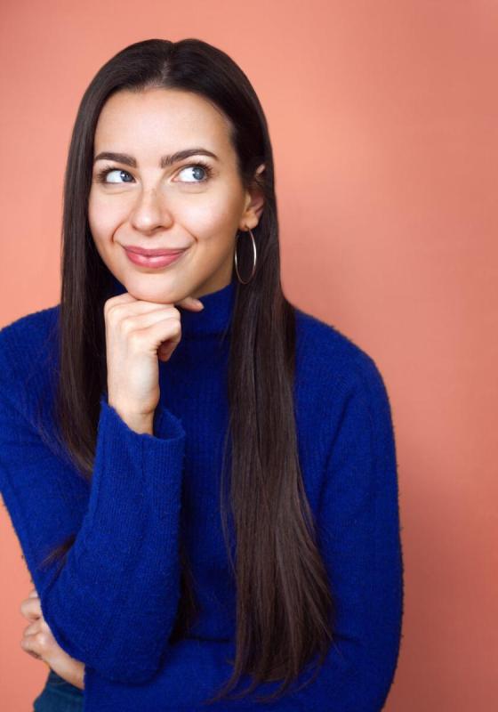 Abi wears a blue roll neck jumper and stands against a muted orange background. They look up towards the sky resting their chin on their fist.