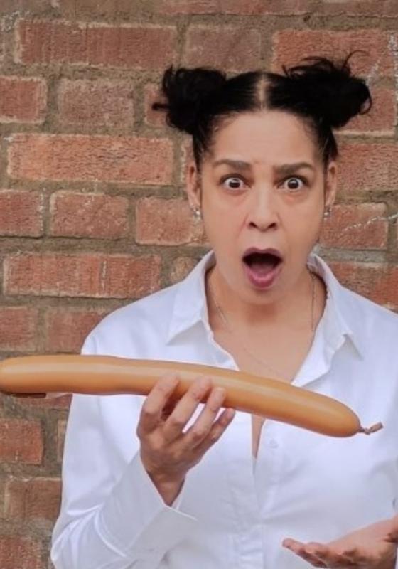 Josephine is wearing a white shirt, standing in front of a brick wall with their jaw dropped. They are holding a long thin balloon. 