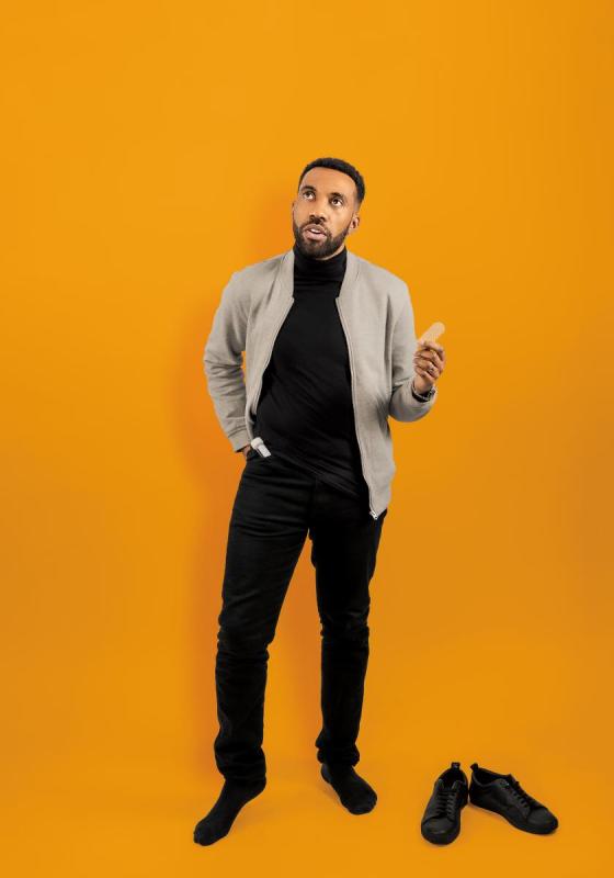 Darran stands against an orange background, one hand in his pocket holding a soggy biscuit. He stands with his shoes off.