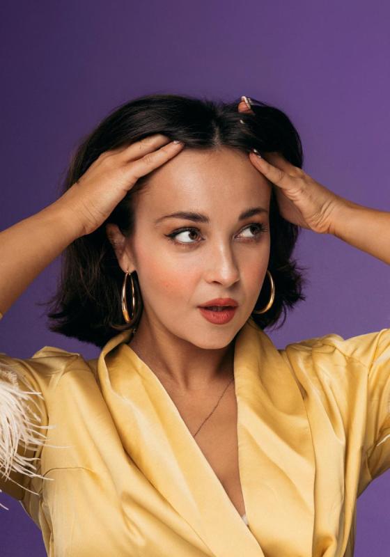 Against a purple background, Sarah Roberts pushes their hair back with their hands. They are wearing a yellow silk shirt with feathered cuffs half out of shop and hoop earrings.