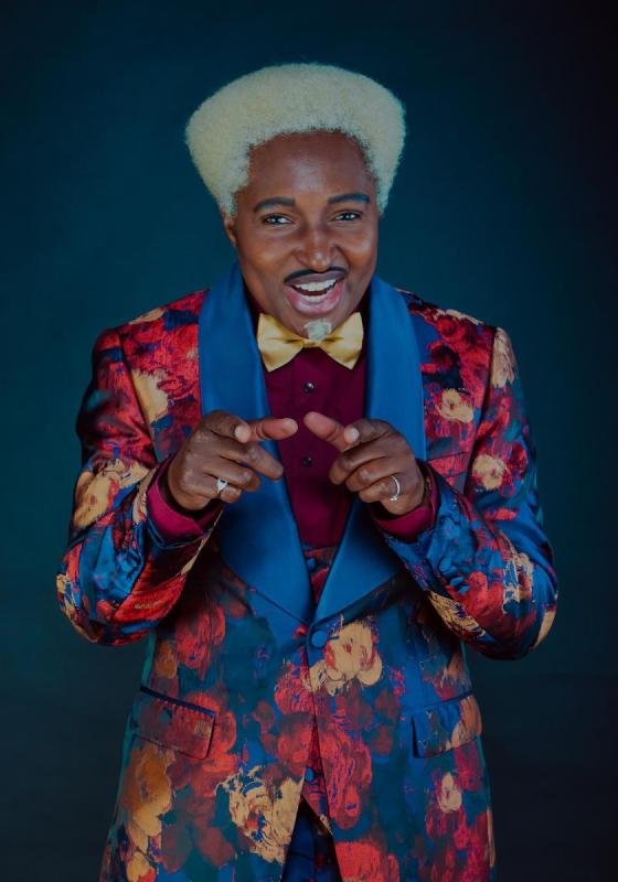 Drag King Don One wears a brightly coloured yellow, red and blue floral suit. They stand in a typical primetime TV host pose with hands cocked, ready to welcome audience members.