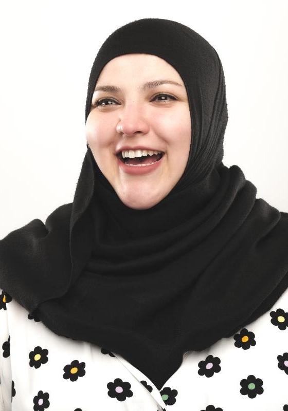 Fatiha wears a black hijab and flower patterned shirt. They are smiling openly to the viewer.