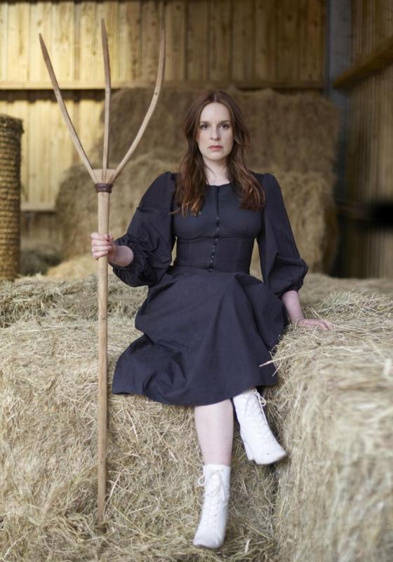 Katie sits with her legs crossed on a bale of hay holding a pitchfork