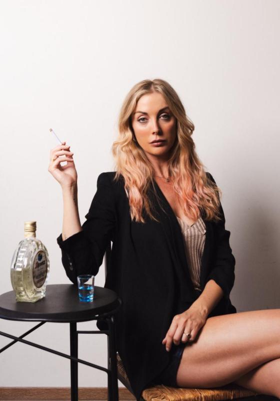 Milanka is sat looking seriously at the camera. She is holding a cigarette and leaning her elbow on a table with a shot glass and bottle of liquer on it. she is wearing a black blazer, short black skirt and a brown top.