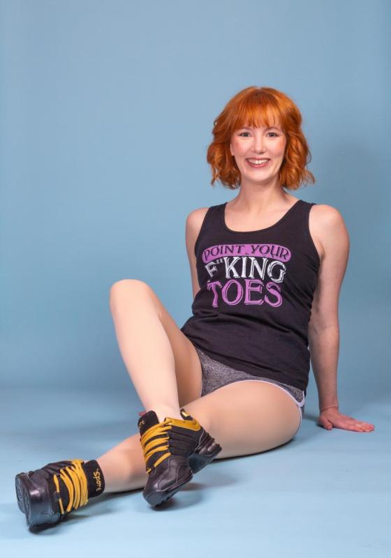 A woman sits on the floor in front of a blue backdrop. She has her legs stretched out with one bent over another. She is smiling innocently but has a top that says 'point your f**king toes'