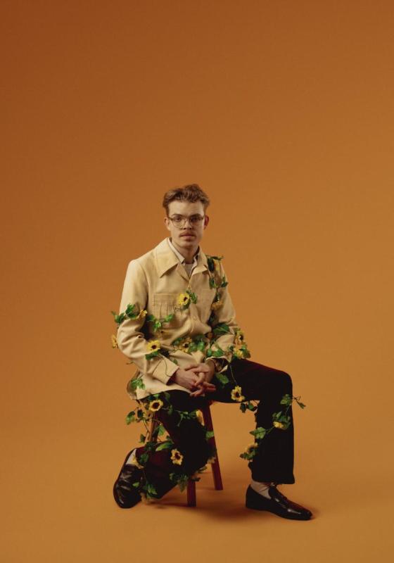 Horation is sat on a chair in front of an orange background. He is wearing a camel coloured jacket, black tailored trousers and black loafers. There is a vine of sunflowers wrapped around his body.