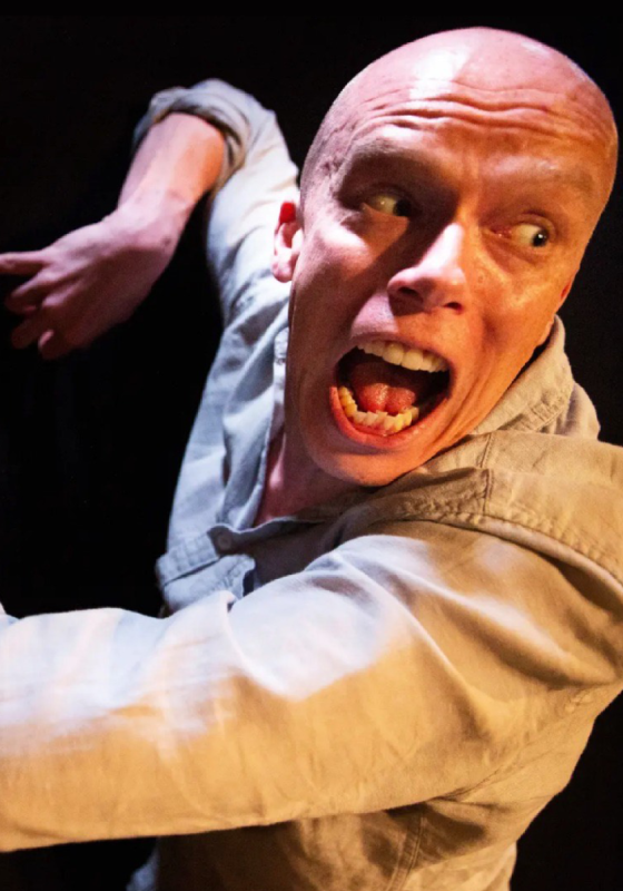 Jordan Brookes' face is close the camera with his eyes wide and mouth open. He wears a beige shirt. His arms are behind him, with his hands tensed.  