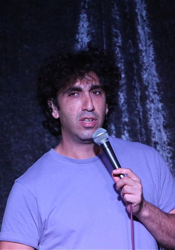 Ray stands in front of a blue velvet backcloth holding a microphone in their right hand wearing a light purple t shirt.