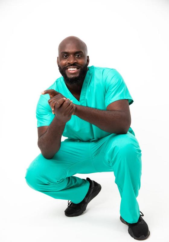 Man in bright blue scrubs crouches and holds his hands together looking at camera