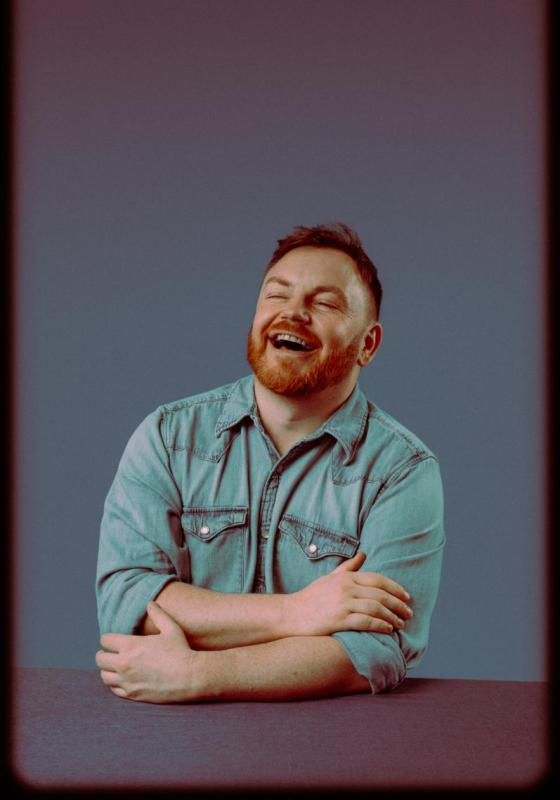 A photo of John, smiling, with eyes closed and arms loosely folded. John wears a light blue pastel denim shirt and has striking red hair. 