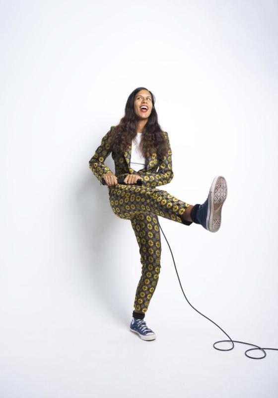 Woman stands in front of white back drop in jazzy suit holding a microphone and extending the right leg