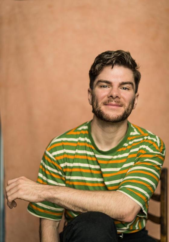Photo of Christopher wearing a horizontally striped orange, cream and green t-shirt. They look relaxed and are sat on a chair with their arm loosely crossed over their knee.