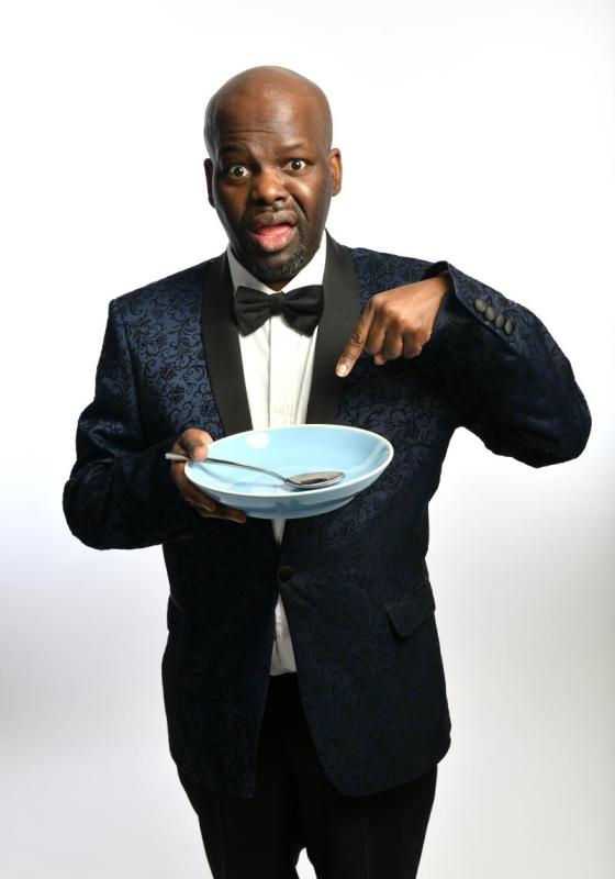 Daliso wears a patterned suit dinner jacket and bow tie. They stand holding an empty bowl and spoon.