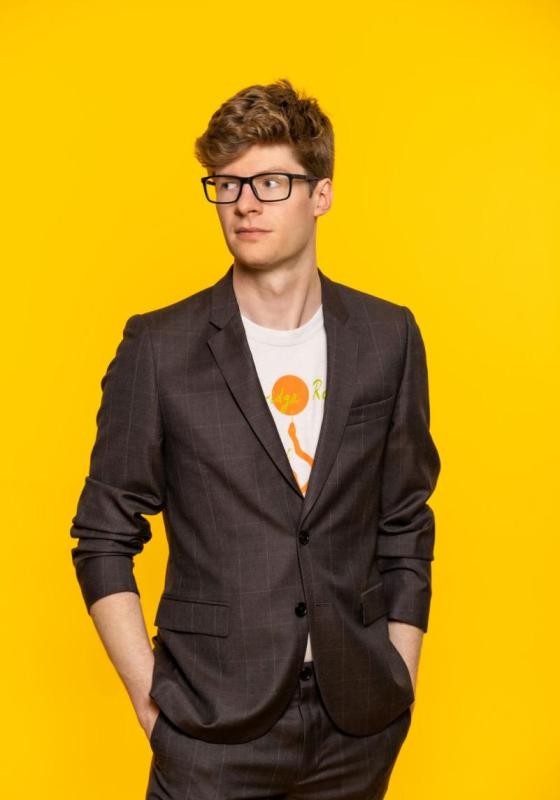 Man in suit stands with hands in pockets, looking to the left in front of yellow background