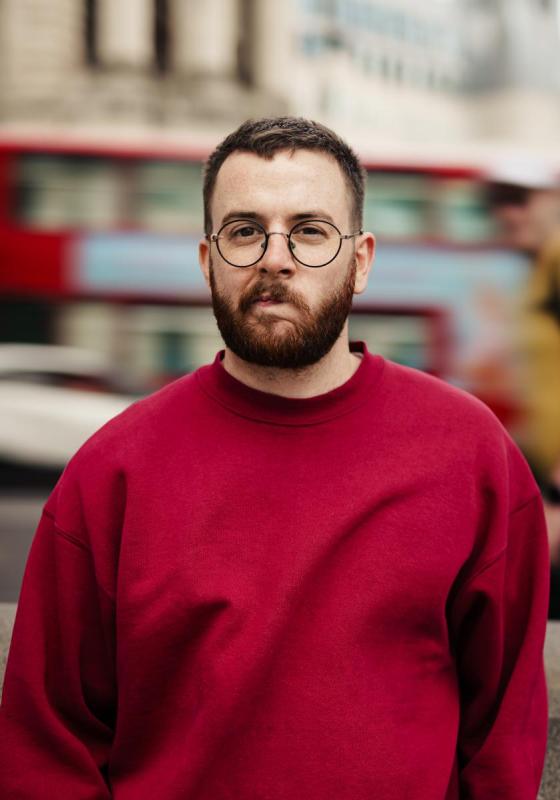 Headshot of Rich wearing a red jumper and round glasses in front of a blurred London bus cityscape background.