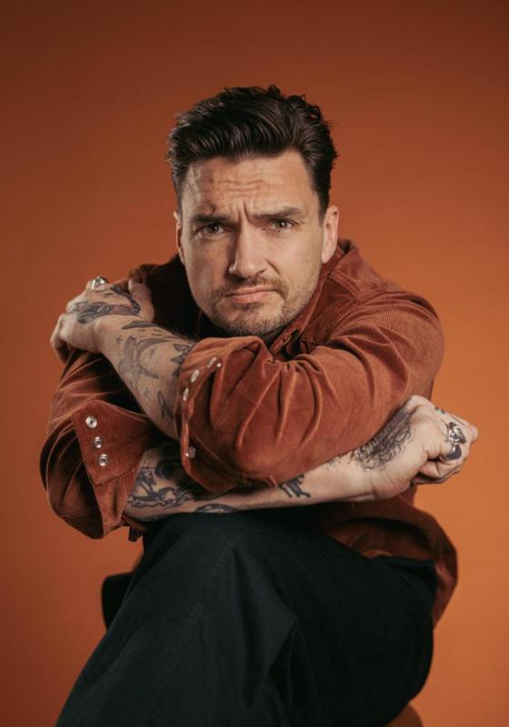 Man sits in front of orange backdrop with arms crossed round himself perched on top of his knee which is bent over his other leg