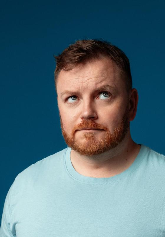 Photo of John looking uncertain with eyes towards the sky. They are wearing a light blue t-shirt against a dark blue teal background.
