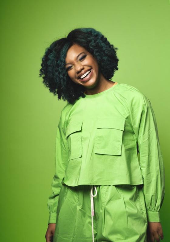 Kemah wears a utilitarian all green long sleeve top and trousers against a background colour of the exact same shade of green.