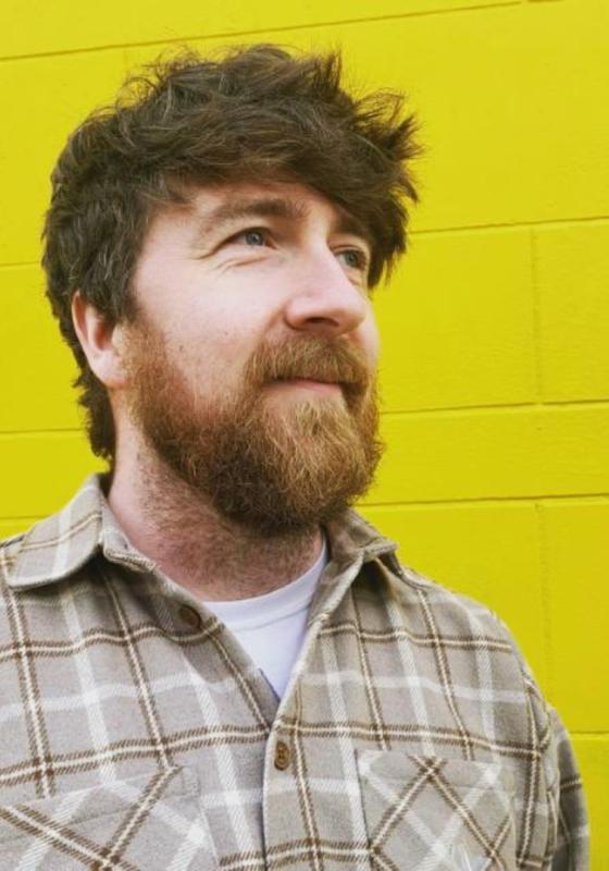 Dan stands against a yellow brick wall wearing a brown checkered shirt.