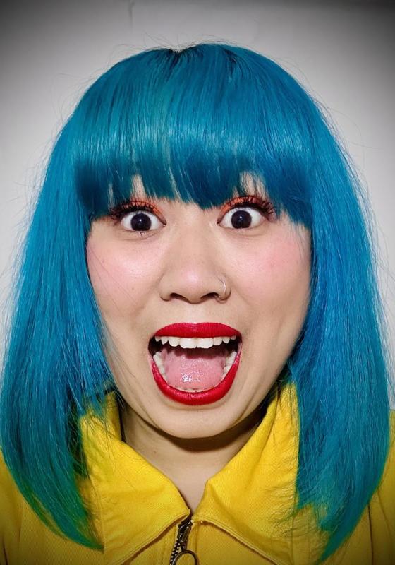 A close-up of the performer's face, whose mouth is open and eyes are wide. She has blue hair and a bright yellow blouse.