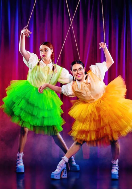 Maddy and Marina are dressed in brightly coloured tutu skirts with platform heels. Their hands are suspended by puppet strings.