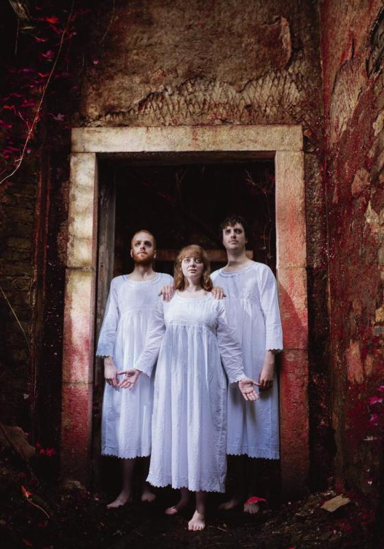 The Tarot trio stand in front of a red stained gothic entrance way in white tunic dresses.