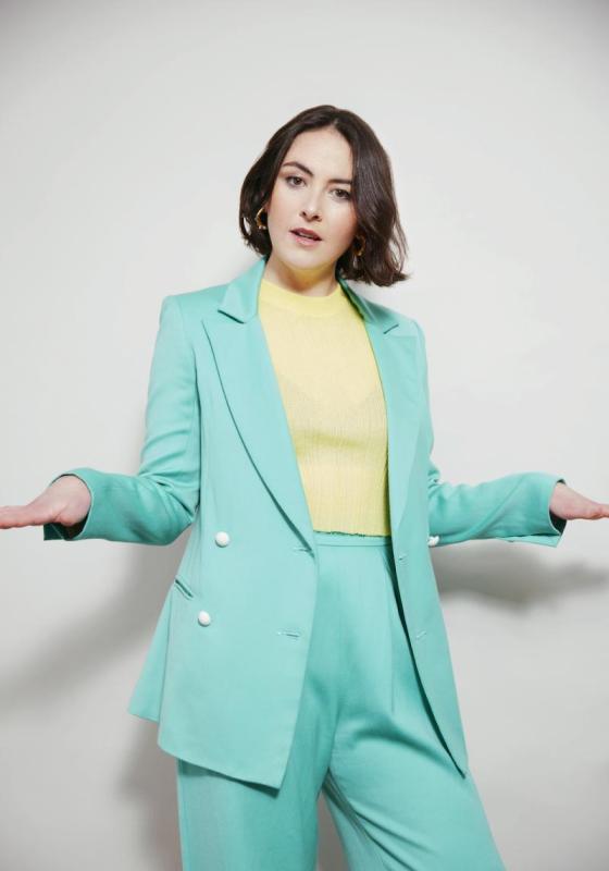 Photo of Emma Sidi wearing a bright blue-turquoise suit and pastel yellow jumper. her hands are outstretched to the sides with palms down.