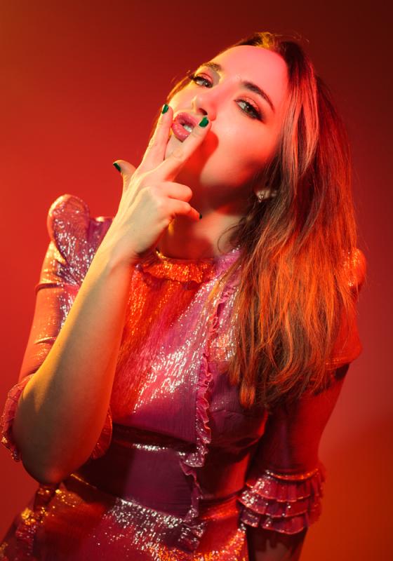 Extremely red-oriented photo with saturation, backdrop and outfit choice. Fern is looking at the camera with two fingers pointed in a suggestive way over her lips.