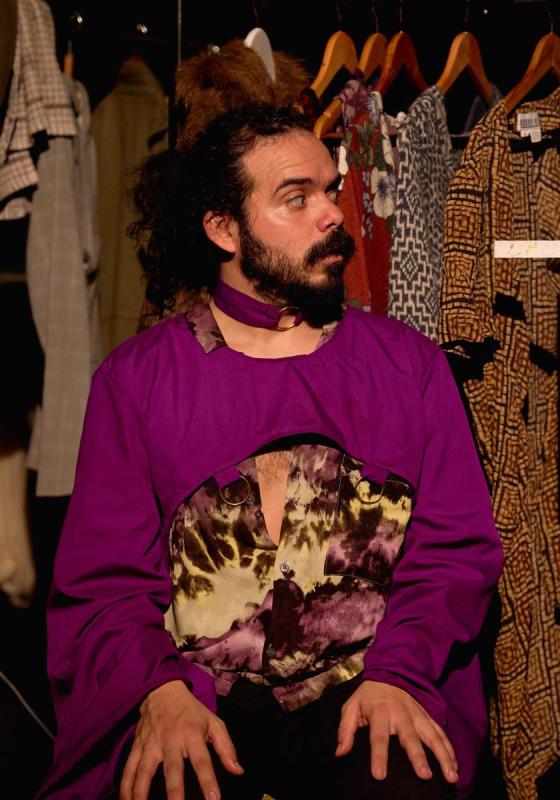Production shot from Jeezus F*cking Christ. Sergio wears a purple robe sitting down in front of a wardrobe rail. A floating hand appears to their right holding a wooden crucifix.