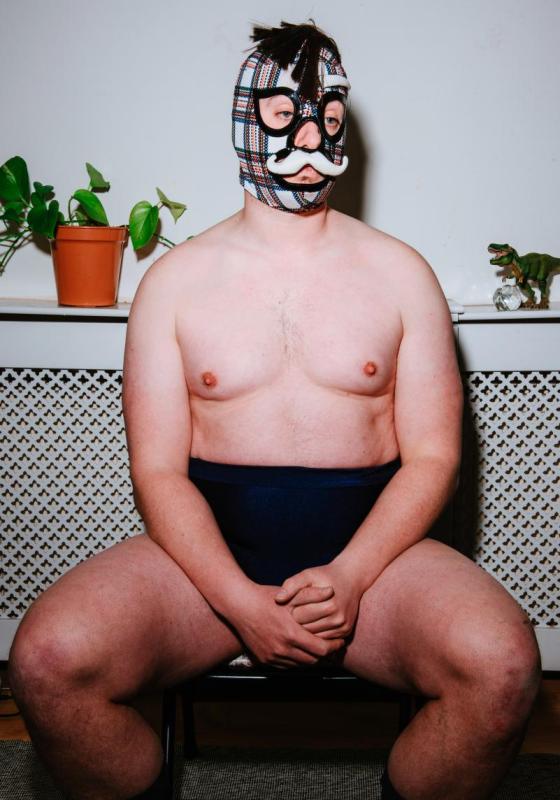 A white man is sat wearing just short, dark navy coloured shorts. He is wearing a checked wrestling mask with a white moustache sown on. He is sat on a chair and on a ledge behind him there is a pot plant and a toy dinosaur. 