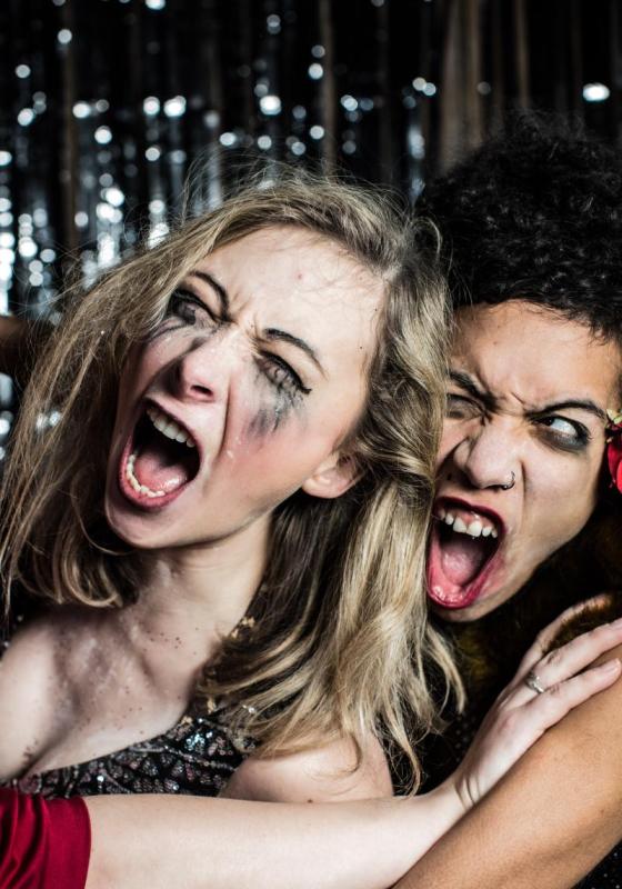 Against a glitter curtain, two women with smeared make-up appear to be fighting and locked in struggle.