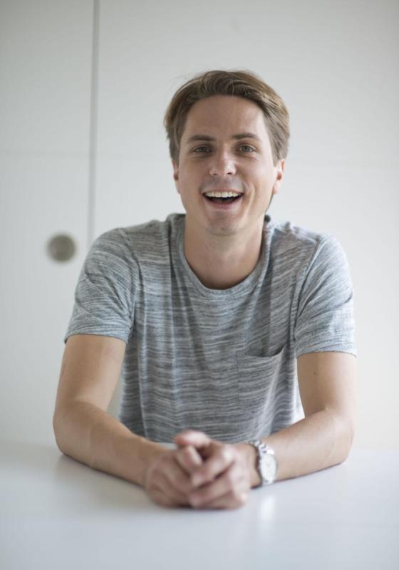 A smiling picture of Joe Thomas wearing a faded subtly grey striped t-shirt and a watch on their left wrist. 
